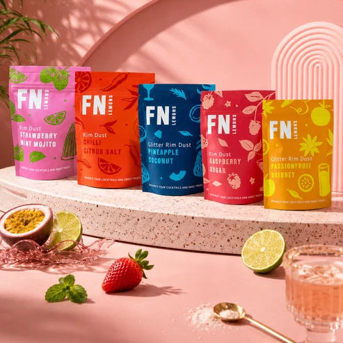 Five colorful pouches of FN Lemons Flavoured Cocktail Rim Salt and sugars arranged on a stone pedestal against a pink studio backdrop, with fruit and a glass of pink drink as styled props.